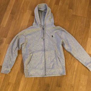 Grå hoodie från Polo Ralph Lauren M - Snygg ljusgrå hoodie från Polo Ralph Lauren med dragkedja framtill, huva med snörning och klassisk broderad logga på bröstet. Tillverkad i mjuk bomullsblandning och har två fickor framtill. Perfekt för chill dagar eller när du vill ha en clean look. Storlek M. Riktigt skön och eftertraktad hoodie som passar bra till allt. säljer då den inte passar mig när jag köpte. Helt oanvänd!