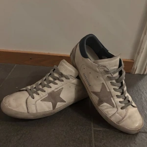 Golden Goose  - Golden Goose sneakers i vitt skinn med grå mockadetaljer och ikonisk stjärna på sidan. Skorna har grå snörning, mörkblå hälkappa och perforerad tunga. Klassisk låg modell med platt sula och cool vintagekänsla. Kommer med originalkartong och dustbag. Ny pris 6500