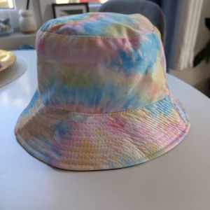 Pastellfärgad och svart bucket hat - Vändbar bucket hat. Ena i pastelliga tie-dye färger med blått, rosa, gult och grönt, och ena helsvart. klassisk form med bred brätte och är tillverkade i bomull. Perfekta för att lyfta din sommarstil.