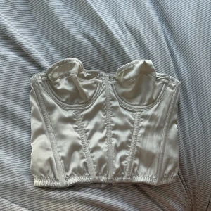 Vit satin korsett från Urban Outfitters - Säljer en snygg vit korsett från Urban Outfitters i satinliknande material. Toppen är axelbandslös med markerade kupor och har dragkedja bak för enkel påklädning. Perfekt att styla med jeans eller kjol för en trendig look. Toppen har några slitningar från förra ägaren ( se sista bild ). 