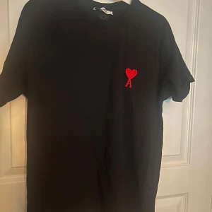 Svart t-shirt från Ami Paris med rött hjärta - Svart t-shirt från Ami Paris med klassisk passform och korta ärmar. På bröstet finns ett broderat rött hjärta med bokstaven A. Tillverkad i mjuk bomull som känns skön mot huden. Perfekt för en clean och stilren look.
