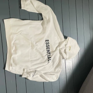 Vit Essentials hoodie XL - Säljer en clean vit hoodie från Essentials i storlek XL. Tröjan har svart tryck på bröstet och ärmen, stor huva och en klassisk magficka. Perfekt för dig som gillar streetwear och vill ha en enkel men snygg look.