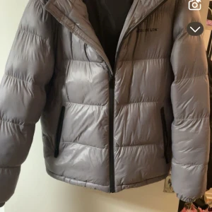 Grå pufferjacka från Closure LDN - Snygg grå pufferjacka från Closure LDN med svarta detaljer och dragkedja framtill. Jackan har en klassisk quiltad look och två fickor med dragkedja. Perfekt för kyliga dagar när du vill hålla stilen.