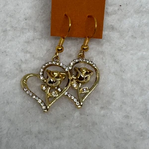 Guldiga örhängen med hjärtan och rosor - Snygga guldiga örhängen med hjärtformad design och en ros i mitten. Kanter dekorerade med glittrande stenar som ger extra bling. Perfekt för dig som gillar romantiska detaljer och vill sticka ut. Krokarna är i sterling silver, nya