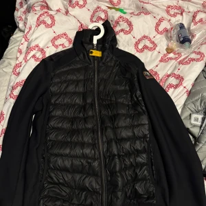 Svart pufferjacka från Parajumpers - Svart pufferjacka från Parajumpers med quiltad front och släta ärmar. Jackan har hög krage, dragkedja och den klassiska gula detaljen vid halsen. Materialmix med vadderad kropp och stickade sidor ger en sportig vibe. Perfekt för kyliga dagar.sen är det lite sönder där nere som ni ser på bilderna