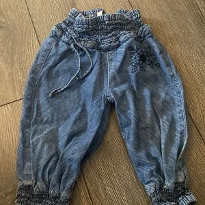 Blå byxor med broderi från KappAhl - Säljer ett par blå byxor från KappAhl med smock i midjan och benslut, snörning i midjan och ett snyggt mörkblått blombroderi på ena benet. Byxorna är i mjukt material och har avslappnad passform, perfekta för lek och rörelse.