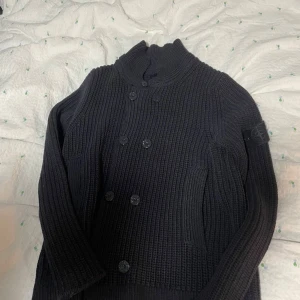 Marin blå  stickad cardigan från Stone Island - Säljer en marin blå, grovstickad cardigan från Stone Island med dubbelknäppning framtill och hög krage. Klassisk patch på ärmen och snygga detaljer med knappar. Det är en extremt sällsynt cardigan och du kommer inte se många med en likadan. Perfekt nu för hösten och vintern! Pris kan diskuteras vid snabb affär. Passar på folket mellan 180-187 ungefär.
