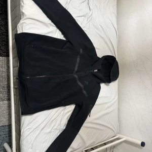 Svart Nike tech Kofta  - Svart hoodiejacka från Nike med dragkedja framtill och huva. Jackan har diskreta grå detaljer och en sportig look. Perfekt för dig som gillar enkel och stilren design.