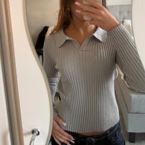 Ljusgrå ribbad långärmad tröja - Säljer en ljusgrå ribbad tröja med lång ärm och snygg krage. Tröjan har en klassisk, lite loose passform och är perfekt för dig som gillar stilrena basplagg. Materialet är mjukt och stretchigt, vilket gör den superbekväm att bära. 