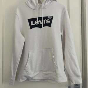 Vit hoodie från Levi's med logga - Vit hoodie från Levi's i klassisk modell med svart logga framtill. Tröjan har huva med snörning, känguruficka och långa ärmar. Tillverkad i mjuk bomullsmix som är perfekt för kyligare dagar. Enkel och stilren design som funkar till det mesta. Har lite små fläckar 