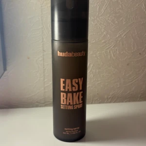 Huda Beauty Easy Bake Setting Spray - En svart, matt flaska med roséguldfärgad text från Huda Beauty. Easy Bake Setting Spray innehåller risstärkelse, niacinamid och hyaluronsyra. Alkoholfri och parfymfri formula, 100 ml. Perfekt för att fixera sminket hela dagen.