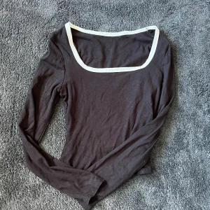 Svart långärmad topp med vit kant - En svart långärmad topp med snyggt fyrkantigt halsringning och vit kontrastkant runt halsen. Tillverkad i mjukt bomullsmaterial, perfekt för en stilren och enkel look. Passar dig som gillar minimalistisk stil och vill ha något bekvämt men ändå trendigt.