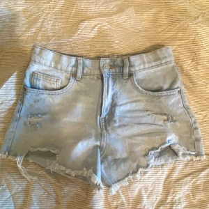 Ljusblå jeansshorts från Zara - Säljer ett par ljusblå jeansshorts från Zara med hög midja, råa fransiga kanter och slitna detaljer framtill. Klassisk femficksmodell med bälteshällor och knappstängning. Perfekta för varma dagar och en avslappnad look.