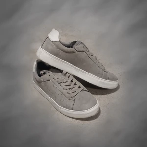 Grå skor med vit detalj - Snygga sneakers i ljusgrå mocka med vit häl och vit sula. Skorna har klassisk låg profil, rund tå och snörning framtill. Perfekta för dig som gillar stilrena och minimalistiska sneakers med en modern touch.