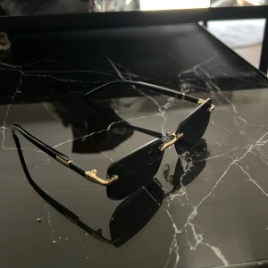 Svarta rektangulära solglasögon med gulddetaljer - Snygga svarta solglasögon med rektangulära linser och tunna guldfärgade detaljer vid bågarna. Glasögonen har minimalistisk design och näskuddar för extra komfort. Perfekt accessoar för att lyfta din stil med en modern och edgy vibe.