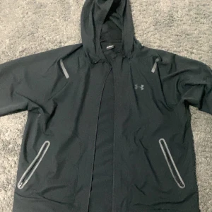 Svart vindjacka från Under Armour XL - Svart vindjacka från Under Armour med huva och grå detaljer vid fickor och axlar.  Finns kvitto och allt 