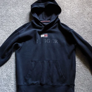 Mörk blå hoodie från Tommy Hilfiger - Mörk blå hoodie från Tommy Hilfiger med klassisk logga och flagga på bröstet. Tröjan har huva, känguruficka och ribbade muddar. Perfekt för dig som gillar stilren streetwear med en touch av premiumkänsla.