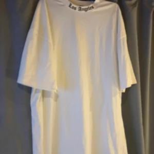 Vit oversized Los Angeles t-shirt - Vit oversized t-shirt med svart Los Angeles-tryck i gotisk stil över hela ryggen och mindre tryck vid halsen framtill. Klassisk rund hals och korta ärmar. Perfekt för en streetwear-look och riktigt skön passform.