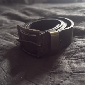 Mocka bälte - Snyggt svart bälte i skinn med klassisk, matt metallspänne där det står 'JEANS The New Fashion'. Bältet har en stilren och rak form med flera hål för justering. Perfekt för att ge jeansen en cool touch.