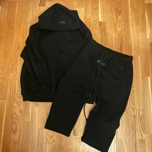 Svart Essentials Fear of God hoodie - Svart hoodie från Essentials Fear of God med stor logga på ryggen och mindre logga på bröstet. Klassisk oversized passform, huva och ribbade muddar. Perfekt för dig som gillar streetwear och vill ha en stilren look.
