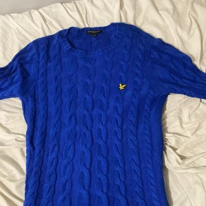 Blå kabelstickad tröja Lyle & Scott - Kabelstickad blå tröja från Lyle & Scott med rund halsringning och gul logga broderad på bröstet. Tröjan är i 100% bomull och har ribbade muddar vid ärmslut och nederkant. Perfekt för dig som gillar klassisk stil med en färgstark twist.