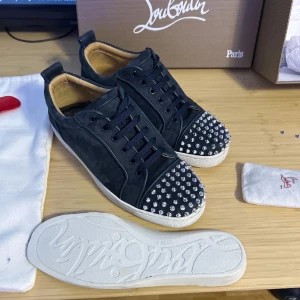 Christian Louboutin svarta sneakers med nitar - Svarta sneakers från Christian Louboutin i mocka med ikoniska röda sulor och silverfärgade nitar på tån. Skorna har svarta snören, vita kanter och en klassisk låg profil. Kommer med extra skosnören, dustbags och originalkartong.