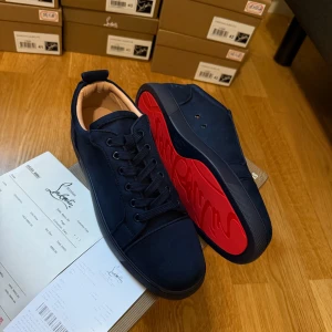 Mörkblå sneakers från Christian Louboutin storlek 42 - Snygga mörkblå sneakers från Christian Louboutin med klassisk röd sula och broderad logga på hälen. Skorna har rund tå, platt sula och är gjorda i mocka med skinnfoder. Perfekt för dig som vill sticka ut med exklusiv stil och detaljer som snörning och diskret branding.