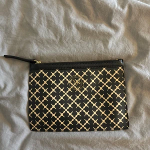 Svart och beige kuvertväska från Malene birger  - Kuvertväska från malene birger i svart och beige med snyggt grafiskt mönster och guldfärgad logga framtill. Väskan har dragkedja upptill och är gjord i tåligt skinnliknande material. Perfekt storlek för det viktigaste och enkel att bära i handen.