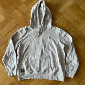 Ljusgrå/beige hoodie från Morris - Hoodie med huva och snörning från Ldrags. Tröjan har en liten svart broderad Morris logga på bröstet. Perfekt för chill dagar och enkel att matcha.