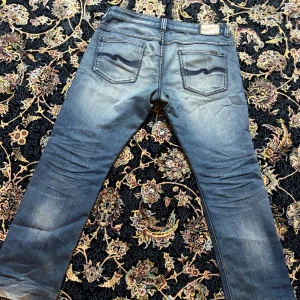 Nudie Jeans blå denim - Nudie Jeans i klassisk blå denim med snygga slitningar framtill och bakfickor med den typiska Nudie-sömmen. Jeansen har en stilren och tidlös look som passar till det mesta. Storlek: W34 L33
