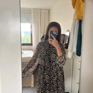 Blommig svart kortklänning från Mango - Supersöt svart kortklänning från Mango med vita och gula blommor. Klänningen har långärmade, lätt puffiga ärmar och en lös passform. Tillverkad i tunn polyester med rund hals och volangdetalj vid kragen. Perfekt för dig som gillar en lekfull och trendig stil.