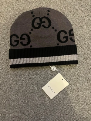 Grå och svart mössa från Gucci - Snygg grå mössa från Gucci med svart GG-logga och ribbad kant i svart och grått. Klassisk design med tydlig logotyp och mjukt material som passar perfekt till höst och vinter. Mössan har en modern och stilren look.
