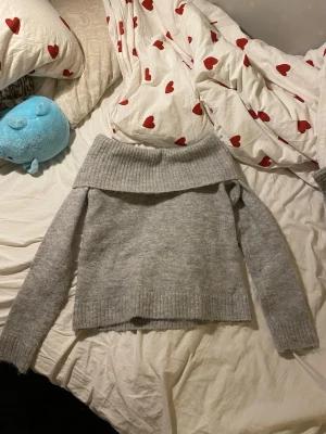 Grå off shoulder stickad tröja Gina Tricot - Mysig grå stickad tröja från Gina Tricot med bred off shoulder-krage och långa ärmar. Tröjan har ribbstickade muddar och är perfekt för lager-på-lager-stil. Enkel att matcha med jeans eller kjol.