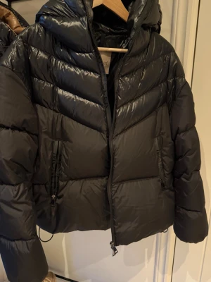 Moncler Svart pufferjacka med huva storlek 3 - Snygg svart pufferjacka från Moncler med stor vadderad huva och dragkedja framtill. Jackan har två sidofickor med dragkedja och en kortare, boxig passform. Materialet är glansigt och quiltat för extra värme och stilpoäng.