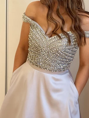 Silverfärgad långklänning med pärlor - Elegant långklänning i silverfärg med offshoulder och v-ringad överdel täckt av pärlor och paljetter. Kjolen är slät och har ett lätt glansigt satintyg som ger en lyxig känsla. Perfekt för bal eller festliga tillfällen.