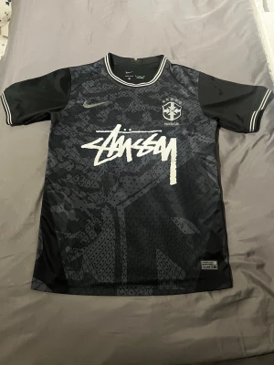 Svart Stüssy x Nike Brasil tröja - Säljer en svart sporttröja från Nike i samarbete med Stüssy och Brasil. Tröjan har ett unikt mönster, vita detaljer och Stüssy-loggan stort framtill. Rund halsringning och korta ärmar med vita ränder. Perfekt för dig som gillar streetwear och sport.
