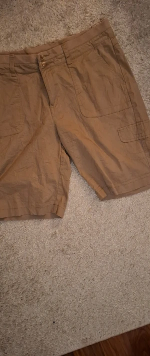 Beige cargoshorts med fickor - Säljer ett par beige cargoshorts med flera praktiska fickor och knappgylf. Shortsen har en avslappnad passform och är perfekta för varma dagar. Tillverkade i ett lätt och bekvämt material som passar till sommaren. Varumärket är Lee