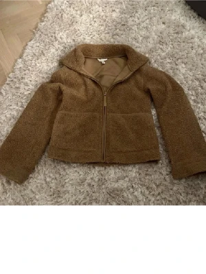 Brun teddyjacka med dragkedja - Mysig brun teddyjacka med dragkedja framtill och stor krage. Jackan har två stora fickor framtill och är tillverkad i ett fluffigt teddy-material som håller dig varm. Perfekt för kyliga dagar och enkel att matcha med olika outfits.