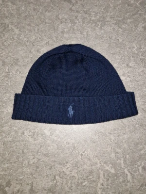 Polo Ralph Lauren Mössa - Mörkblå stickad mössa från Polo Ralph Lauren med klassisk uppvikt kant och broderad logga framtill. Perfekt accessoar för kyliga dagar, gjord av bomull/ull.