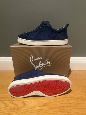 Blå Christian Louboutin sneakers - Snygga mörkblå sneakers från Christian Louboutin i mocka med klassiska röda sulor och coola nitar på tån. Skorna har vita sulor och matchande blå skosnören. Perfekta för dig som vill sticka ut med exklusiv streetstyle.