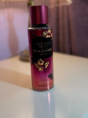 Victoria's Secret Pure Seduction Noir - En doftmist från Victoria's Secret i serien Pure Seduction Noir. Flaskan är cylinderformad och har en snygg mörklila till svart färgskiftning med guldiga blommotiv. Innehåller 250 ml och har en metallisk lila kork. Perfekt för dig som gillar fruktiga och sensuella dofter.