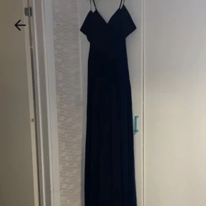 Marinblå långklänning med smala band - Elegant marinblå långklänning med smala axelband och v-ringad urringning. Klänningen har en flytande siluett och är perfekt för speciella tillfällen. Tillverkad i ett mjukt, lätt material som ger ett snyggt fall.
