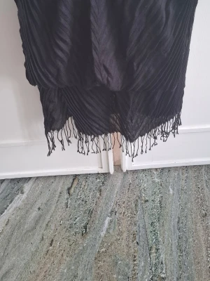 Svart plisserad halsduk med fransar - Snygg svart halsduk med plisserad struktur och tunna fransar längs kanterna. Perfekt accessoar för att lyfta din outfit. Mjuk och lätt i materialet, passar bra till jackor och kappor.