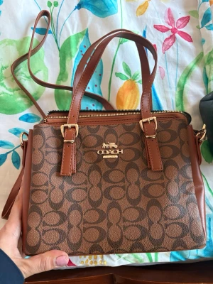 Brun handväska från Coach - Snygg brun handväska från Coach med klassiskt monogrammönster och guldfärgade detaljer. Väskan har både handtag och justerbar axelrem samt flera fack med dragkedja. Tillverkad i konstläder med svart tygfoder inuti. Perfekt för dig som gillar stilrena accessoarer.