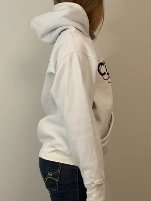 Vit hoodie med CLASSY-tryck - Säljer en vit hoodie från Classy med stor huva och magficka. På bröstet står det 'CLASSY' i svart och rött tryck. Hoodien har en avslappnad passform och ribbade muddar vid ärmar och midja. Perfekt för en chill och stilren look.
