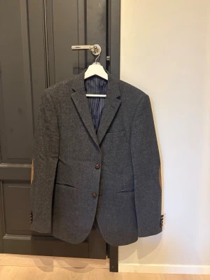 Grå Sand tweedkavaj - Stilren grå tweedkavaj med bruna mockalappar på armbågarna och bruna knappar. Kavajen har klassisk passform, två framfickor och en bröstficka. Fodret är randigt i blått och vitt. Perfekt för en dressad look med en twist.