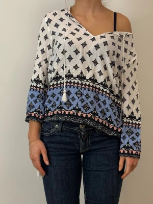 Mönstrad blus med blå och rosa detaljer - Snygg blus med svart mönster på vit botten och blå samt rosa detaljer längs ärmar och nederkant. Blusen har v-ringning, snörning med tofsar och en avslappnad passform. Perfekt för dig som gillar boho-stil och vill ha något unikt i garderoben.