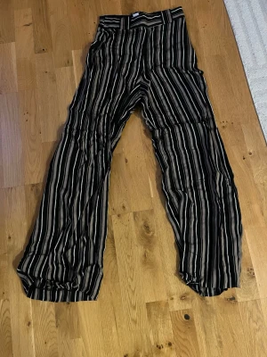 Randiga vida kostymbyxor - Säljer ett par svarta kostymbyxor med vita och beige vertikala ränder. Byxorna har hög midja och vida ben för en avslappnad och trendig look. Perfekta för dig som gillar att sticka ut med mönster och vill ha en bekväm passform.