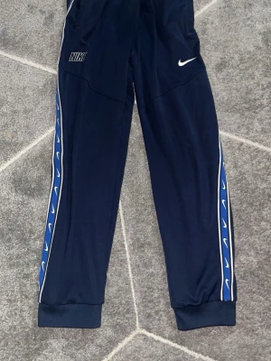 Mörkblå Nike mjukisbyxor med logga - Snygga mörkblå mjukisbyxor från Nike med vita loggor längs sidorna och vita detaljer. Byxorna har resår i midjan och vid bensluten samt sidofickor. Perfekta för chill eller träning och har en sportig look. Det är ett sett samma look på koftan