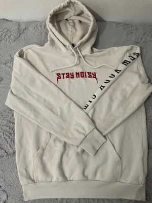 Vit hoodie med tryck Stay Noisy - Vit hoodie med huva och dragsko, röd text 'STAY NOISY' på bröstet och svart tryck längs ena ärmen. Klassisk känguruficka framtill och ribbade muddar. Perfekt för en avslappnad streetwear-look.